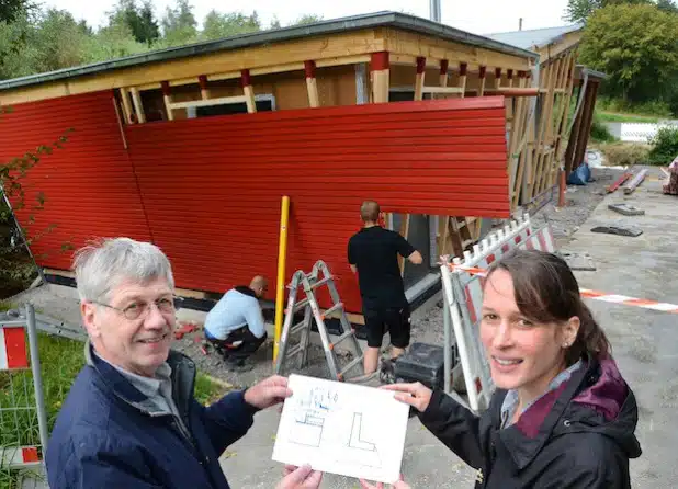 Architekt Johannes-Ulrich Blecke und Projektleiterin Verena Scholten begutachten den Baufortschritt am Waldschiff im Bibertal in Rüthen. Christian Wünsche (hinten l.) und Jonas Hengsbach (hinten r.) von der Firma Blüggel aus Schmallenberg bringen zurzeit die prägnante rote Fassade an (Foto: Thomas Weinstock/Kreis Soest).