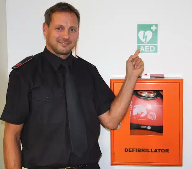 Jan Falke an einem Defibrillator im Olper Kreishaus. Das AED-Piktogramm weist häufig den Weg (Foto: Kreis Olpe).